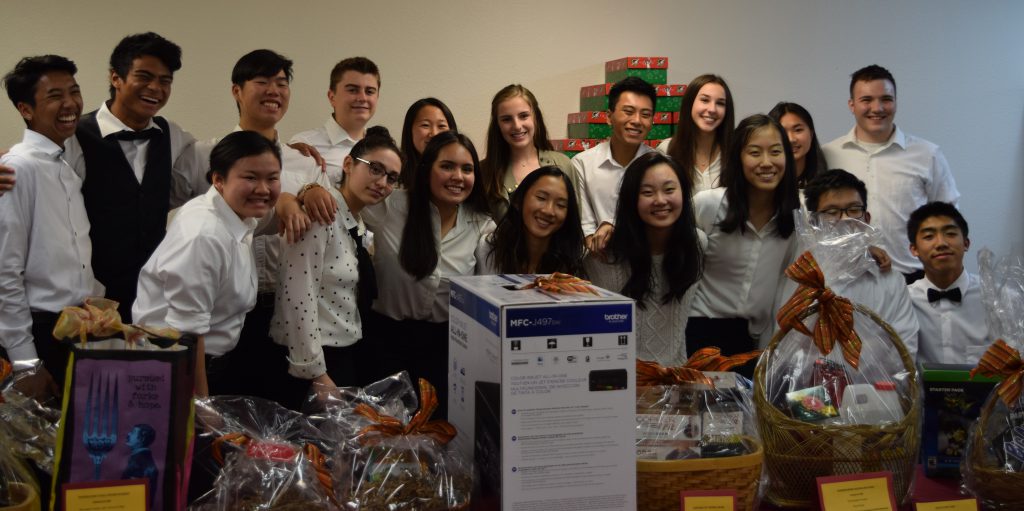 A group of teenage Key Club officers, both girls and boys, dressed in white tops and black trousers posing for a picture in front of silent auction items.