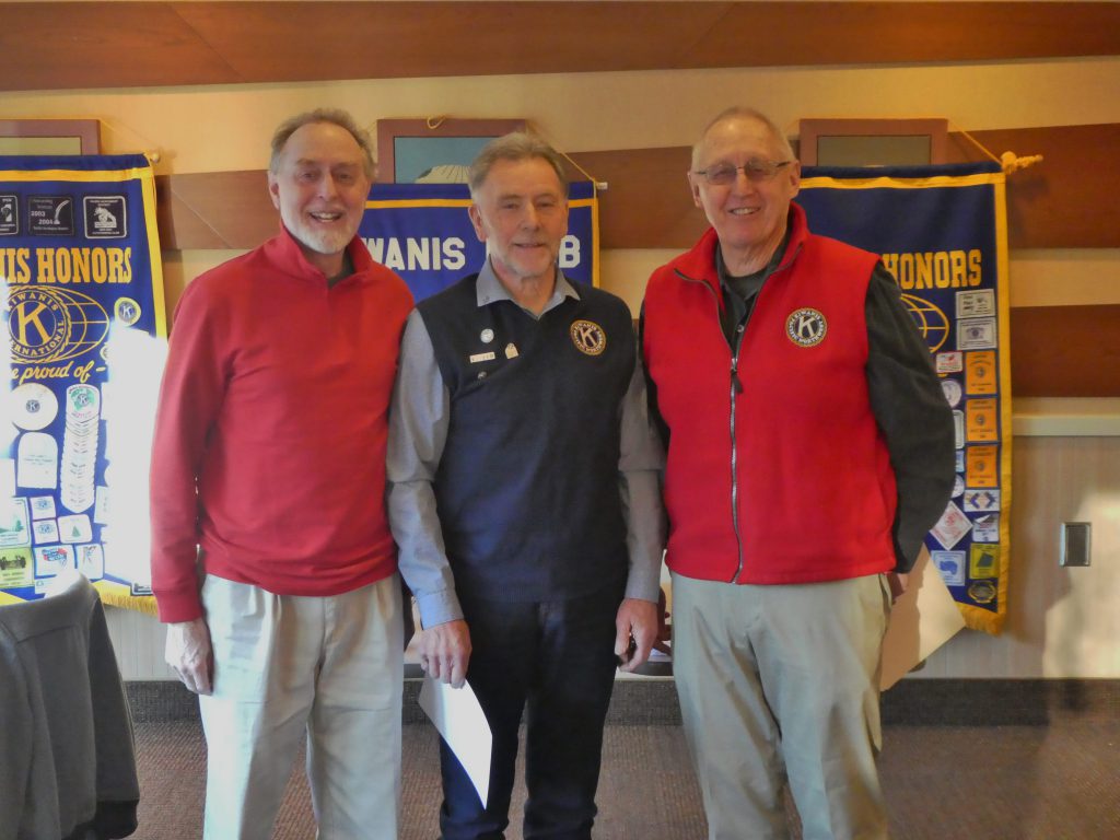 Two smiling men in red tops flanking a third gentleman in a navy blue vest. Background consists of three Kiwanis banners,