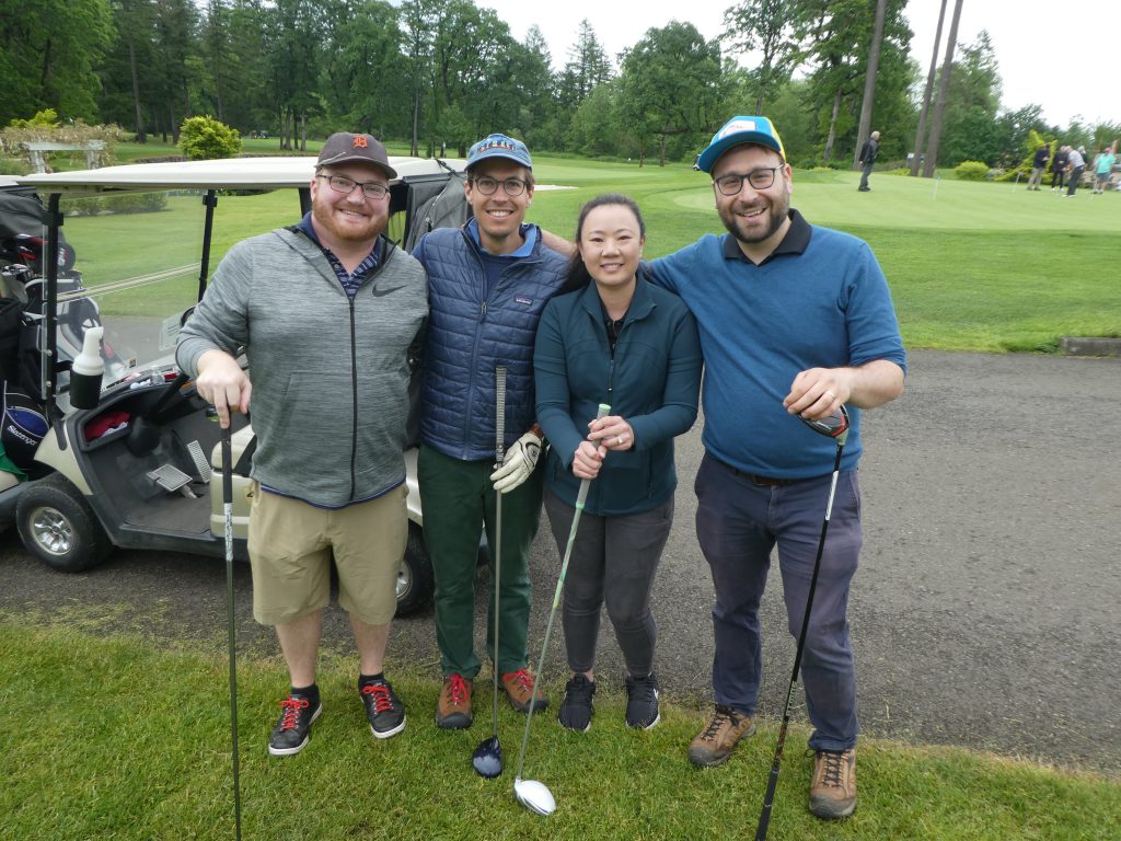 Three male and one female golfer holding golf clubs and standing in front of a golf cart with at Camas Meadows Golf Course.