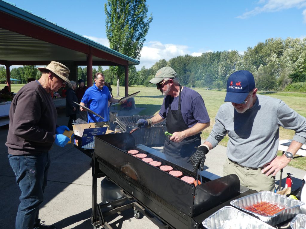 Four members in a sunny park setting working over a giant grill as they cook burgers for over 200 people gathered nearby.