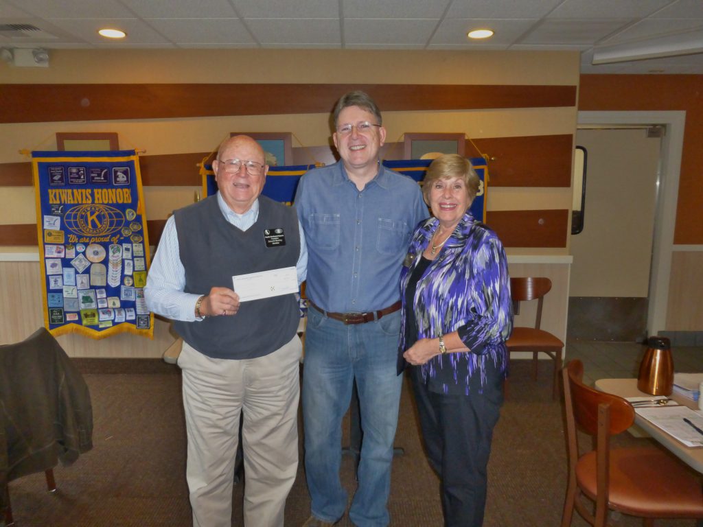 Two men dressed in blue, one holding a check, and one woman in a purple patterned jacket. Background consists of tables and Kiwanis Honors banners.